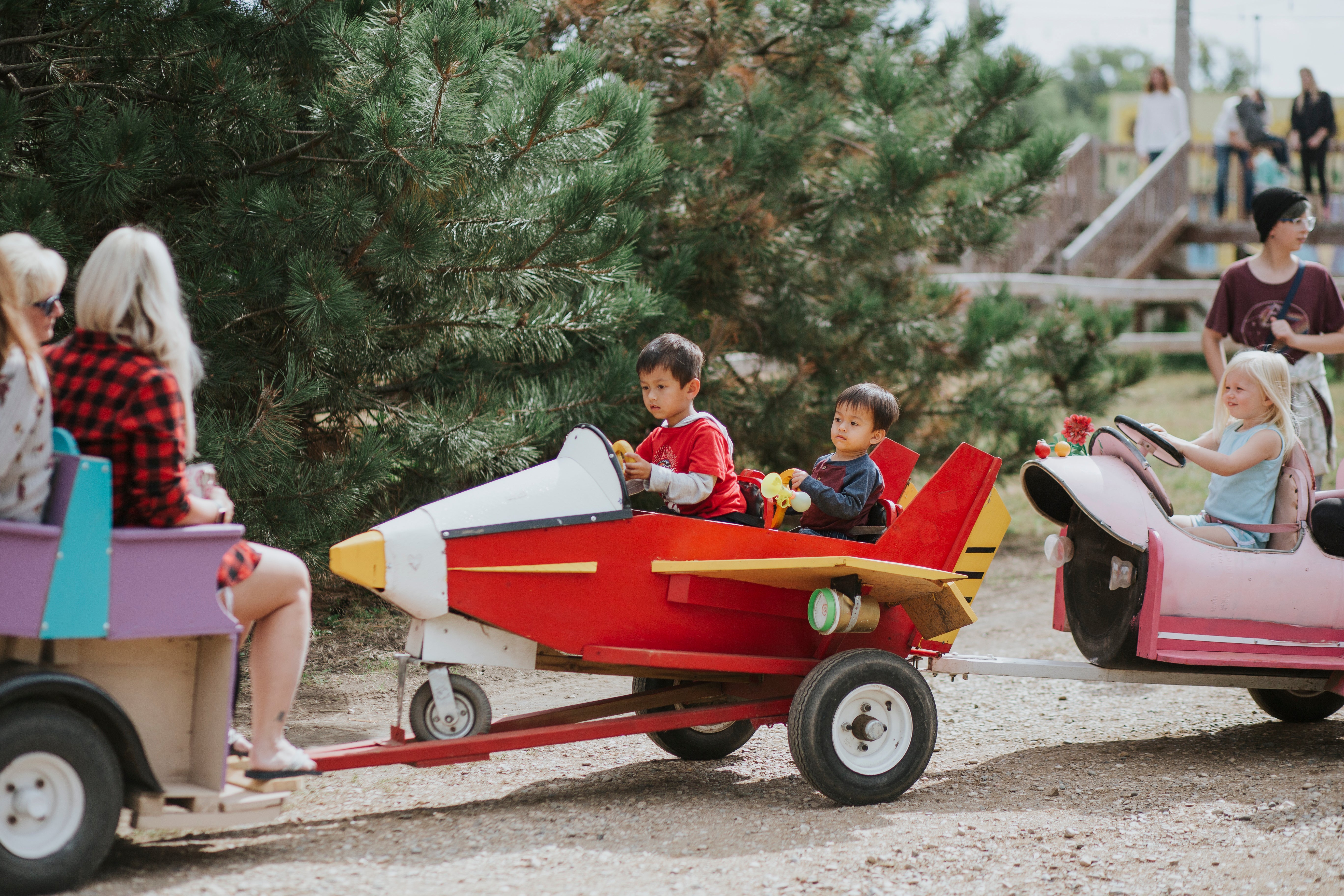 A family outdoor scene at a rustic park or fair, featuring children driving small toy vehicles. In the center, a young boy in a red shirt drives a red and yellow rocket-shaped cart being pulled by a woman in a red plaid shirt.